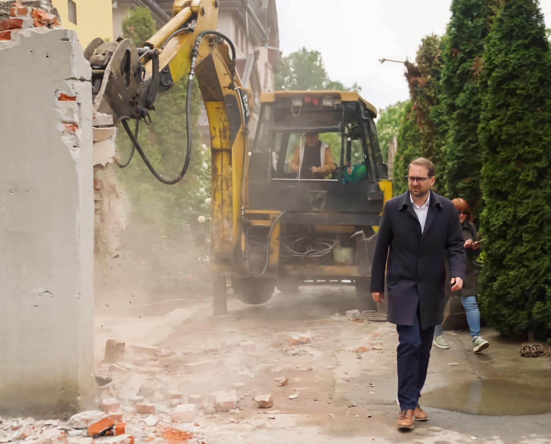 Dominic Fritz, în palton închis la culoare, trecând pe lângă un excavator care dărâmă un zid, cu praf și moloz pe jos