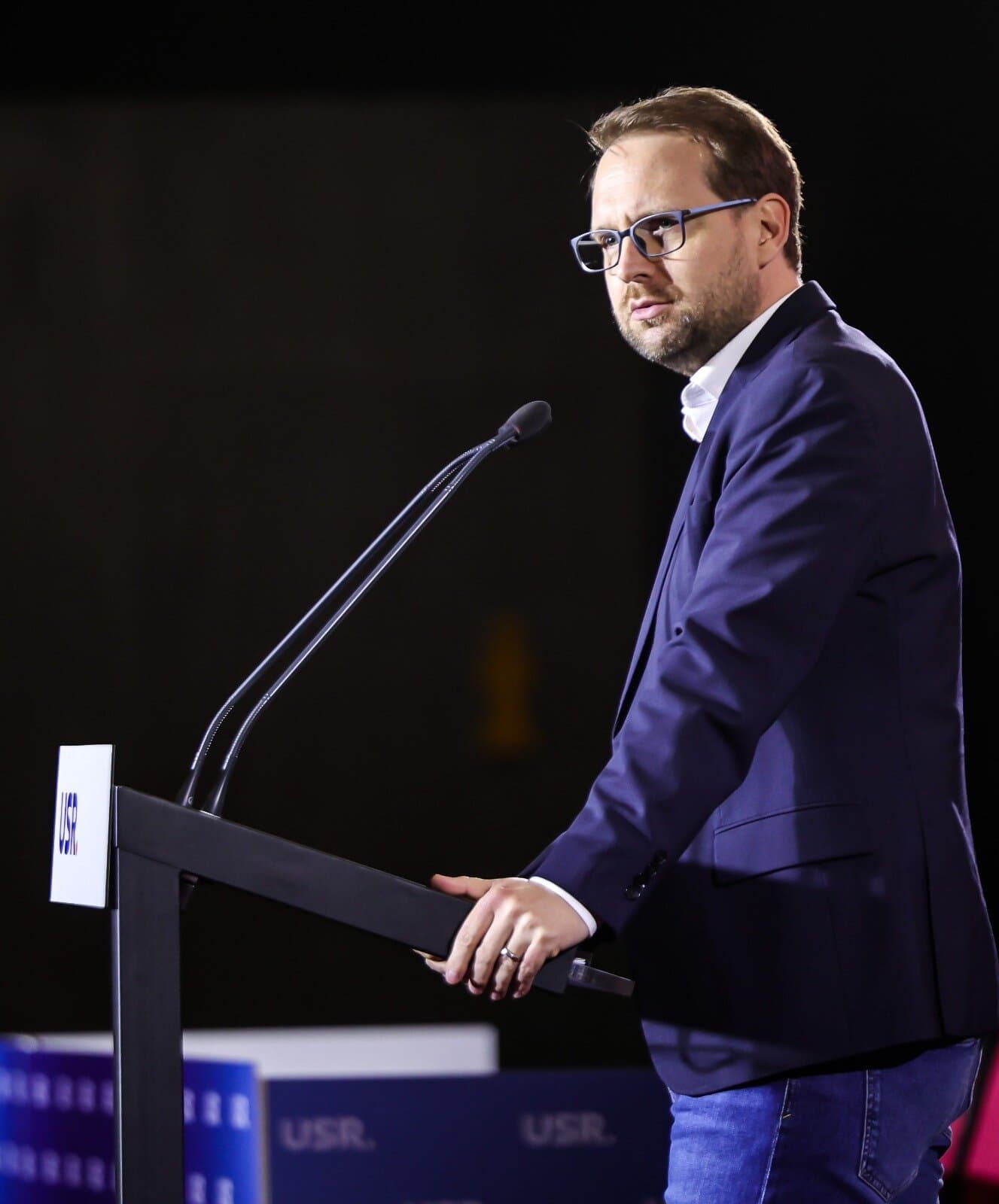 Dominic Fritz speaking from a lectern marked with the USR logo, lit against a dark stage